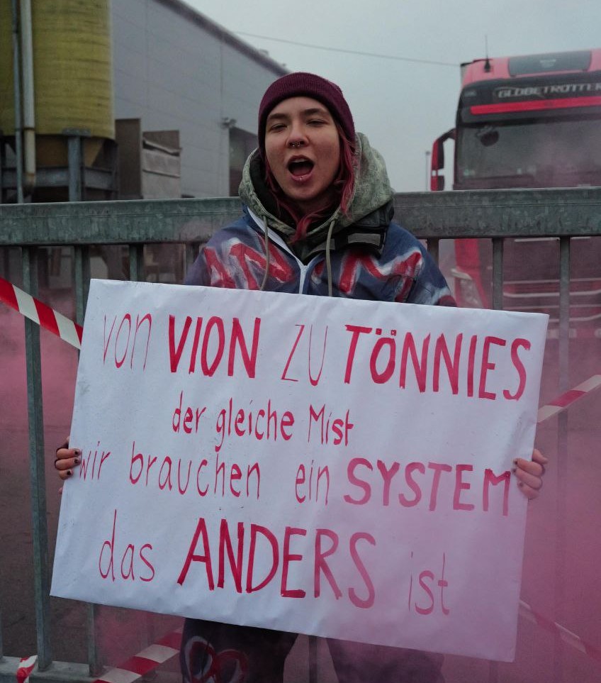 Eine Aktivistin steht vor einem verschlossenen Tor und hält ein Schild von dem Steht "Von Vion zu Tönnies der gleich Mist - Wir brauchen ein System das Anders ist". Im Hintergrund wartet ein LKW vor dem Tor.