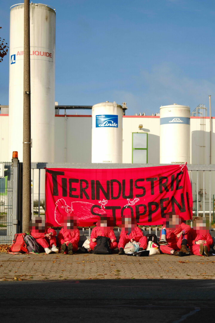 Menschen blockieren die Einfahrt einer Fabrik, im Hintergrund hängt ein Transparent mit der Aufschrift Tierindustrie stoppen